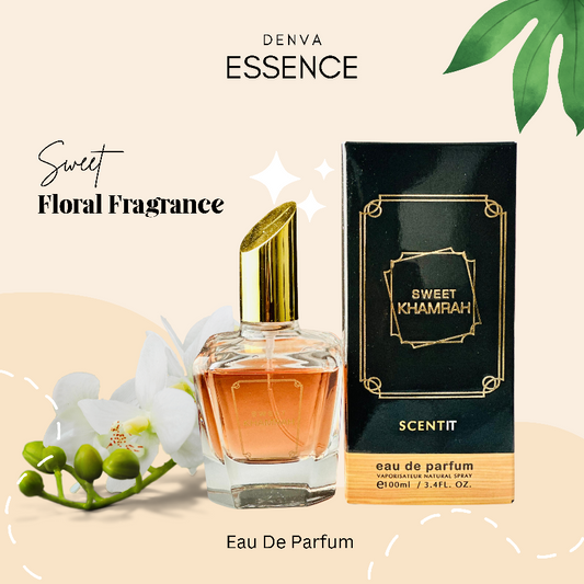 A product shot of Denva Essence Sweet Khamrah perfume. The bottle, filled with a light amber liquid and topped with a gold cap, is positioned next to its black and gold box. A sprig of white flowers with green buds is placed near the bottle. The background is a light gradient with stylized green leaves in the upper corners. Text on the image includes "DENVA ESSENCE," "Sweet Floral Fragrance," "SWEET KHAMRAH," "SCENTIT," "eau de parfum," "VAPORISATEUR NATURAL SPRAY," and "100ml/3.4FL. OZ."